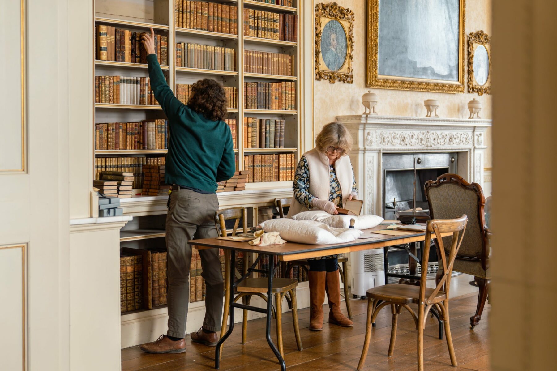 A man removes books from a shelf whilst a woman inspects them on a table in a library at Powderham Castle