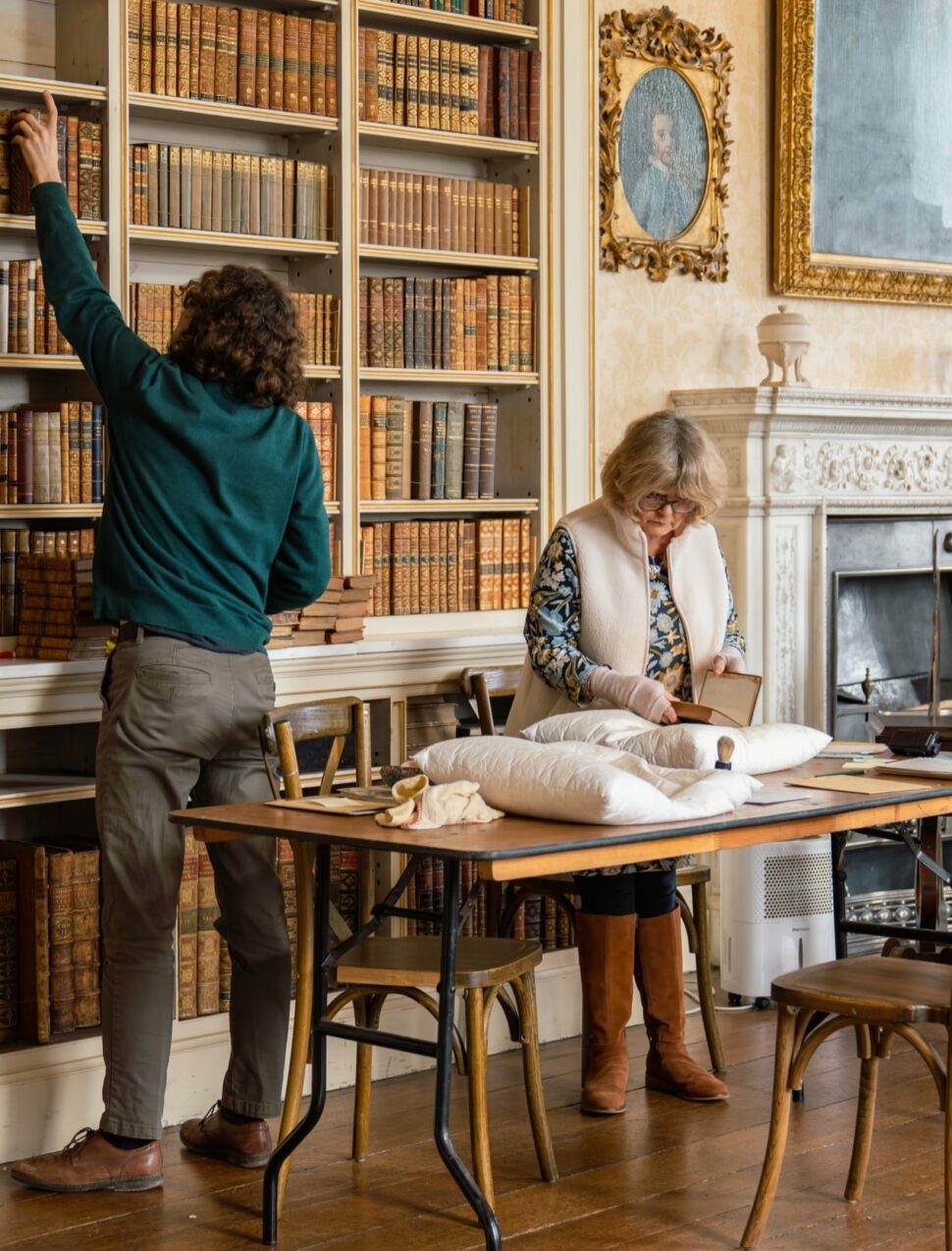 A man removes books from a shelf whilst a woman inspects them on a table in a library at Powderham Castle